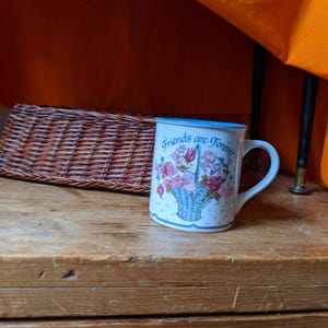 May include: White ceramic mug with a blue rim and handle. The mug features a floral basket design with pink and red roses and the text "Friends are Forever". A decorative item for the kitchen or a gift for a friend.