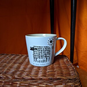 May include: A pale blue ceramic mug with a white handle. The mug features a black and white sheep illustration with the text "A ewe called Sue". A sun and floral design are also present.