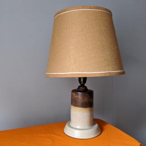 May include: A ceramic table lamp with a tan burlap shade and a white trim. The base is cylindrical, with a gradient of brown and beige tones. The lamp sits on a round, off-white base, creating a warm, inviting aesthetic.