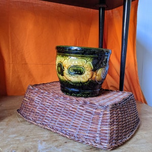 May include: A brown wicker basket with a green and yellow ceramic planter on top. The planter has a floral design.