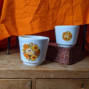 May include: Two white ceramic flower pots with a floral design in shades of orange, yellow, and brown. The pots are sitting on a wooden surface with drawers.
