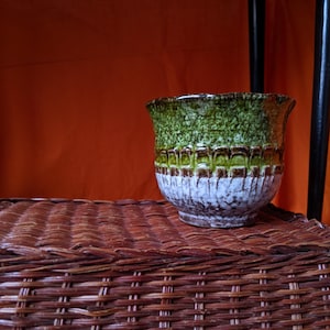 May include: A ceramic vase with a textured surface. The top is green, transitioning to brown and white. The vase has a unique, layered design. The vase is sitting on a brown wicker basket.