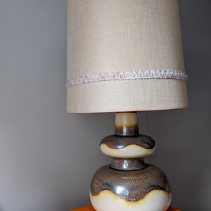 May include: A retro ceramic table lamp with a beige fabric shade. The lamp base features a two-tiered design with a brown and white glaze. A white decorative trim encircles the shade. The lamp sits on an orange surface against a grey wall.