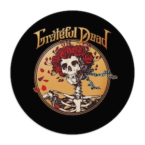 May include: A round black graphic featuring the Grateful Dead logo in gold lettering above a skeleton with a crown of red roses. The skeleton is inside a gold circle, with red rose petals scattered around.