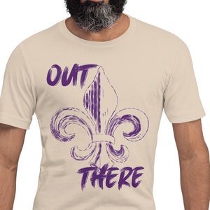 Puede incluir: Una camiseta beige con una flor de lis morada y el texto "OUT THERE" impreso en la parte delantera.