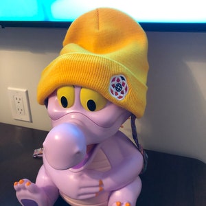 May include: A purple dragon-shaped plastic bucket with a yellow beanie on its head. The beanie has a white logo with a circle and a star inside. The bucket has a purple strap.