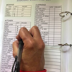 Puede incluir: Una lista de verificaci&oacute;n blanca con una ilustraci&oacute;n en blanco y negro de una camioneta vintage en la parte superior. La lista de verificaci&oacute;n se titula "Everyday Essentials List" y est&aacute; dividida en dos secciones: "Bedding" y "Cleaning". La lista de verificaci&oacute;n incluye art&iacute;culos como almohadas, una colcha, toallas, papel de cocina, toallas de t&eacute;, pa&ntilde;os de cocina y papel higi&eacute;nico. Una mano sostiene una pluma negra y marca los art&iacute;culos de la lista de verificaci&oacute;n.