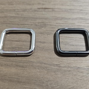 May include: Two square metal rings, one silver and one black, are displayed on a wooden surface. The rings have a hinged design, suggesting they can be opened and closed. These rings are likely used in bag making or other crafting projects.