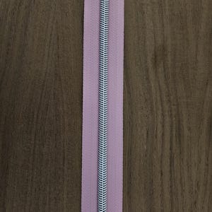 Puede incluir: Primer plano de una cremallera lavanda con un tirador plateado. La cremallera est&aacute; centrada sobre un fondo de madera marr&oacute;n. Los dientes de la cremallera son plateados y la tela es de color lavanda liso.