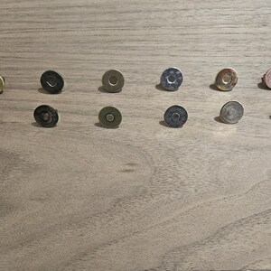 May include: An assortment of magnetic snap closures in various metallic finishes, including gold, silver, and bronze. The snaps are round with a flat, circular design, and are arranged on a wooden surface. These closures are commonly used in bags and other craft projects.