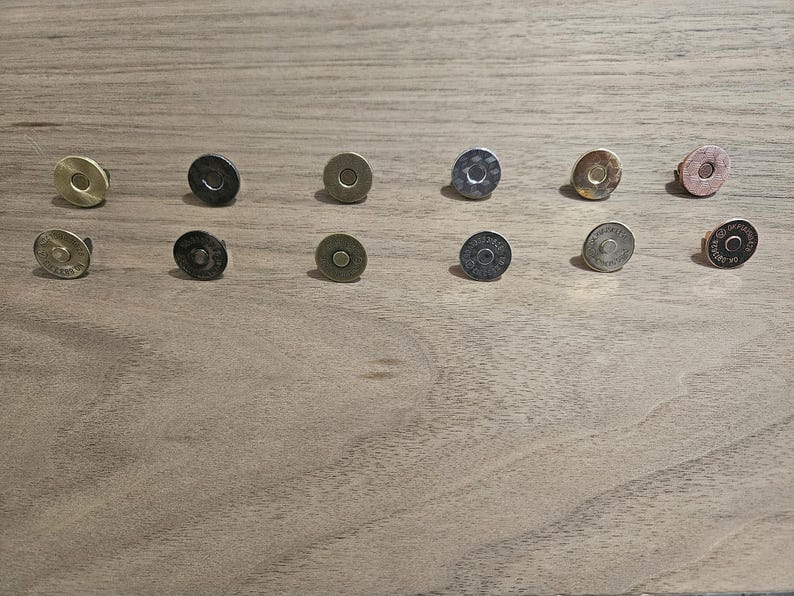 May include: An assortment of magnetic snap closures in various metallic finishes, including gold, silver, and bronze. The snaps are round with a flat, circular design, and are arranged on a wooden surface. These closures are commonly used in bags and other craft projects.