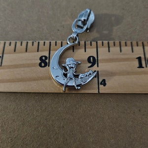 May include: Silver-toned zipper pull featuring a crescent moon design with a seated figure wearing a hat. The zipper pull is attached to a metal zipper. The image is set against a wooden ruler.
