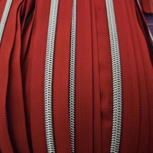 Puede incluir: Primer plano de varias cremalleras rojas con dientes de metal plateado. Las cremalleras est&aacute;n cuidadosamente dispuestas, mostrando el contraste entre la vibrante tela roja y la plata met&aacute;lica. La imagen resalta la textura y el detalle de la construcci&oacute;n de la cremallera.