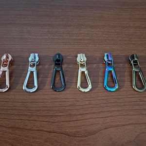 May include: Seven zipper pulls in different colors and finishes, including rose gold, silver, black, gold, iridescent, and bronze. The zipper pulls are all the same shape, a simple oval.