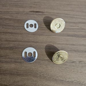 May include: Four metal magnetic snap closures. Two silver-colored, flat, circular pieces with two rectangular holes. Two gold-colored, circular pieces with a raised center and text around the edge. The items are on a brown wooden surface.
