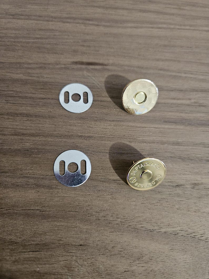 May include: Four metal magnetic snap closures. Two silver-colored, flat, circular pieces with two rectangular holes. Two gold-colored, circular pieces with a raised center and text around the edge. The items are on a brown wooden surface.