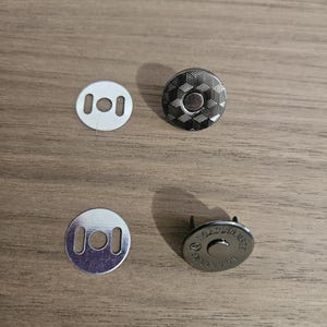 May include: Four pieces of metal hardware for crafting. Two silver-colored, round, flat pieces with two rectangular holes each. Two magnetic snap closures with a geometric pattern and a central circle.