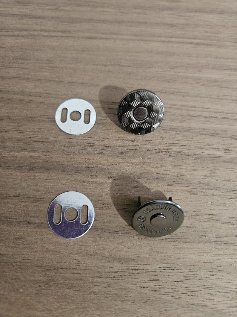 May include: Four pieces of metal hardware for crafting. Two silver-colored, round, flat pieces with two rectangular holes each. Two magnetic snap closures with a geometric pattern and a central circle.