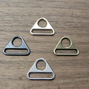 May include: Four triangular metal hardware connectors arranged in a diamond pattern. Each piece features a circular top opening and a rectangular bottom slot. The set includes one gold-tone, one gunmetal, one antique brass, and one silver-tone finish.