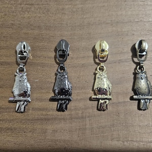 May include: Four metal zipper pulls with owl designs. The pulls are silver, black, gold, and bronze. Each owl is perched on a branch. The zipper pulls are displayed on a wooden surface.