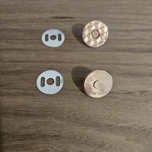 May include: Four magnetic snap closures for crafts. Two silver-colored, rectangular plates with three holes each, and two round, rose-gold colored magnetic snaps. One snap has a textured, diamond pattern, and the other has stamped text.