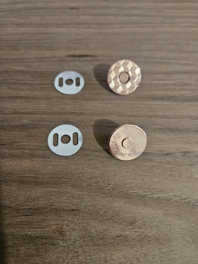 May include: Four magnetic snap closures for crafts. Two silver-colored, rectangular plates with three holes each, and two round, rose-gold colored magnetic snaps. One snap has a textured, diamond pattern, and the other has stamped text.