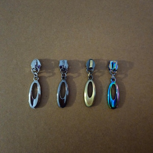 May include: Four metal zipper pulls in silver, dark silver, gold, and iridescent finishes. Each pull has an oval-shaped design with a central cutout and a small chain attached to the top. The pulls are displayed against a brown background.