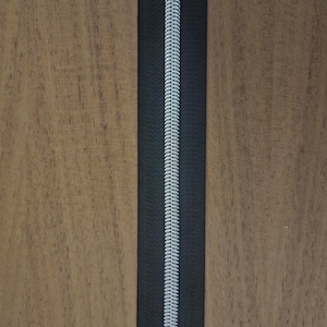 Puede incluir: Primer plano de una cremallera negra con una espiral plateada. La cremallera est&aacute; centrada verticalmente sobre un fondo de madera. Los dientes de la cremallera est&aacute;n entrelazados, creando una l&iacute;nea continua.