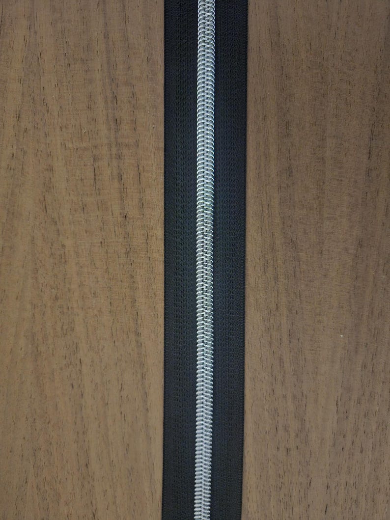 Puede incluir: Primer plano de una cremallera negra con una espiral plateada. La cremallera est&aacute; centrada verticalmente sobre un fondo de madera. Los dientes de la cremallera est&aacute;n entrelazados, creando una l&iacute;nea continua.