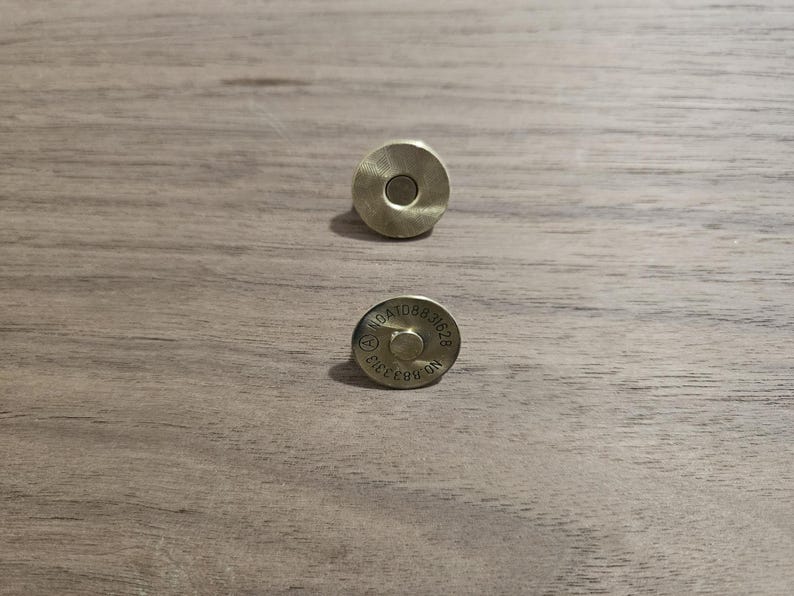 May include: Two brass-colored magnetic snap closures. One is a solid disc with a central hole, the other has stamped text including "NO.883313". These closures are likely for bags or other craft projects.