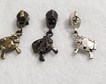 Puede incluir: Tres tiradores de cremallera metálicos con dijes de vaca en bronce, negro y plata. Los dijes de vaca tienen un diseño detallado y están unidos al tirador con un pequeño lazo.