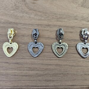 May include: Four metal zipper pulls with heart-shaped charms. One is gold-toned, and the others are silver-toned. Each heart has a decorative border and a cutout heart in the center. The word "Love" is inscribed on the heart.