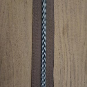 Puede incluir: Primer plano de una cremallera marr&oacute;n con un tirador plateado. La cremallera est&aacute; centrada sobre un fondo de madera. La cinta de la cremallera es de color marr&oacute;n oscuro.