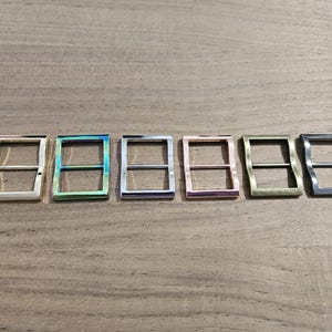 May include: Six metal belt buckles in various colors, including silver, iridescent green, purple, pink, gold, and gunmetal gray. The buckles have a rectangular shape with a wavy design on the outer edges.