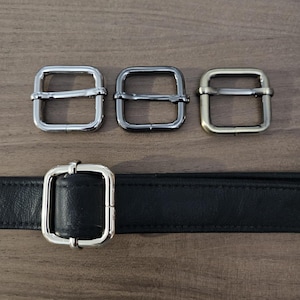 May include: Three metal belt buckle options in silver, black nickel, and antique brass. The buckles are square shaped with a center bar. A black leather belt is shown with a silver buckle.