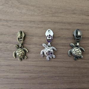 May include: Three metal zipper pulls with turtle charms. The pulls are in antique brass, silver, and antique bronze finishes. The turtle charms have detailed shell patterns and are suspended from the zipper heads.