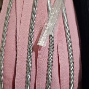 Puede incluir: Primer plano de un rollo de cremalleras rosas con dientes de metal plateado. Las cremalleras est&aacute;n enrolladas, mostrando la tela y los componentes met&aacute;licos. Un paquete de pl&aacute;stico transparente es parcialmente visible, que contiene una cremallera.