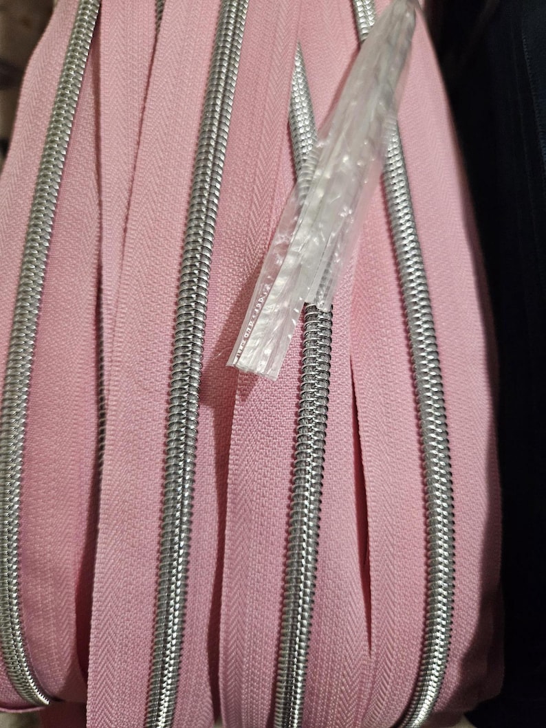Puede incluir: Primer plano de un rollo de cremalleras rosas con dientes de metal plateado. Las cremalleras est&aacute;n enrolladas, mostrando la tela y los componentes met&aacute;licos. Un paquete de pl&aacute;stico transparente es parcialmente visible, que contiene una cremallera.