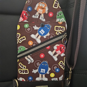May include: A brown crossbody bag with a zippered pocket and a strap. The bag is decorated with a pattern of M&M's characters and the words "Big Fun". The bag has a brown leather bottom with the text "No Sip Butch" printed on it.