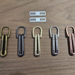May include: An assortment of metal belt buckles in various finishes, including gold, gunmetal, and rose gold. Two matching metal belt keepers are also present. The buckles have a unique design with a hinged mechanism.