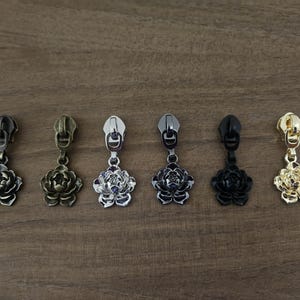 May include: A set of six metal zipper pulls, each featuring a three-dimensional flower charm. The pulls are finished in various metallic tones including antique bronze, brass, silver, gunmetal, matte black, and polished gold, arranged in a row.