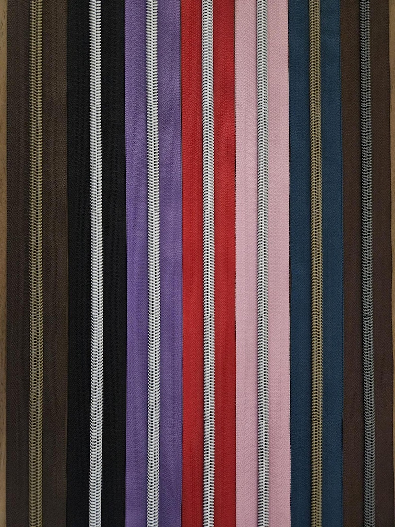 Puede incluir: Una colecci&oacute;n de cremalleras coloridas en varios tonos, incluyendo marr&oacute;n, negro, morado, rojo, rosa y azul. Cada cremallera tiene un tirador de metal contrastante. Las cremalleras est&aacute;n dispuestas verticalmente.