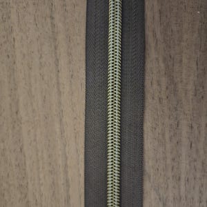 Puede incluir: Primer plano de una cremallera gris oscuro con una espiral de color oro met&aacute;lico. La cremallera est&aacute; centrada sobre una superficie de madera marr&oacute;n claro. Los dientes de la cremallera est&aacute;n entrelazados. Suministros de costura.