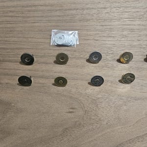 May include: An assortment of magnetic snap closures in various metallic finishes, including gold, bronze, and silver. A small bag of white backing washers is also present. These closures are designed for use in bags, wallets, and other craft projects.
