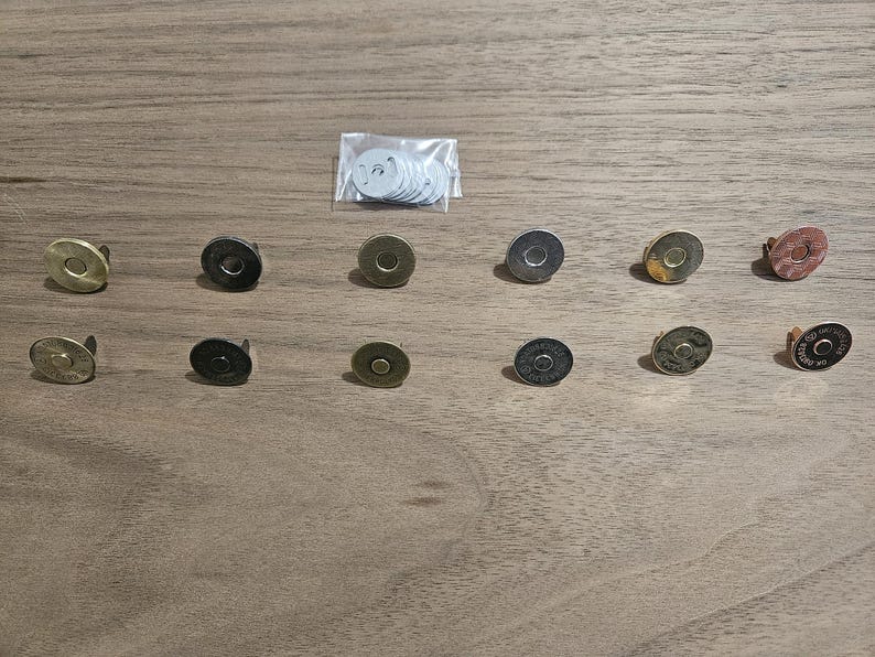 May include: An assortment of magnetic snap closures in various metallic finishes, including gold, bronze, and silver. A small bag of white backing washers is also present. These closures are designed for use in bags, wallets, and other craft projects.
