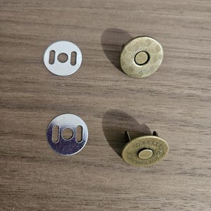 May include: Four magnetic snap closures for bags. Two silver-colored, round, flat pieces with two rectangular holes each. Two bronze-colored, round pieces with a textured surface and a central stud. One piece has the number "883310" printed on it.
