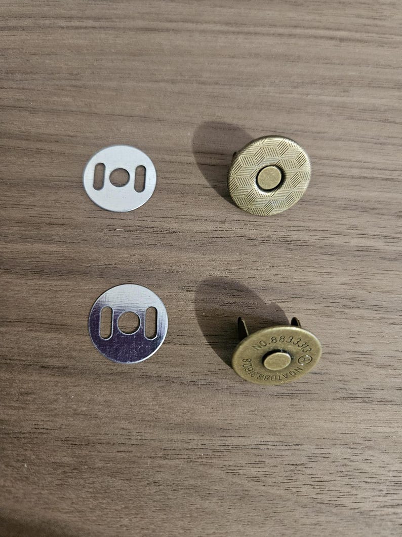 May include: Four magnetic snap closures for bags. Two silver-colored, round, flat pieces with two rectangular holes each. Two bronze-colored, round pieces with a textured surface and a central stud. One piece has the number "883310" printed on it.