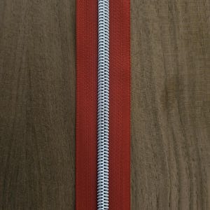 Puede incluir: Primer plano de una cremallera roja con un tirador de metal plateado. La cremallera est&aacute; centrada en una superficie de madera. Los dientes de la cremallera son visibles y la tela es de color rojo liso.