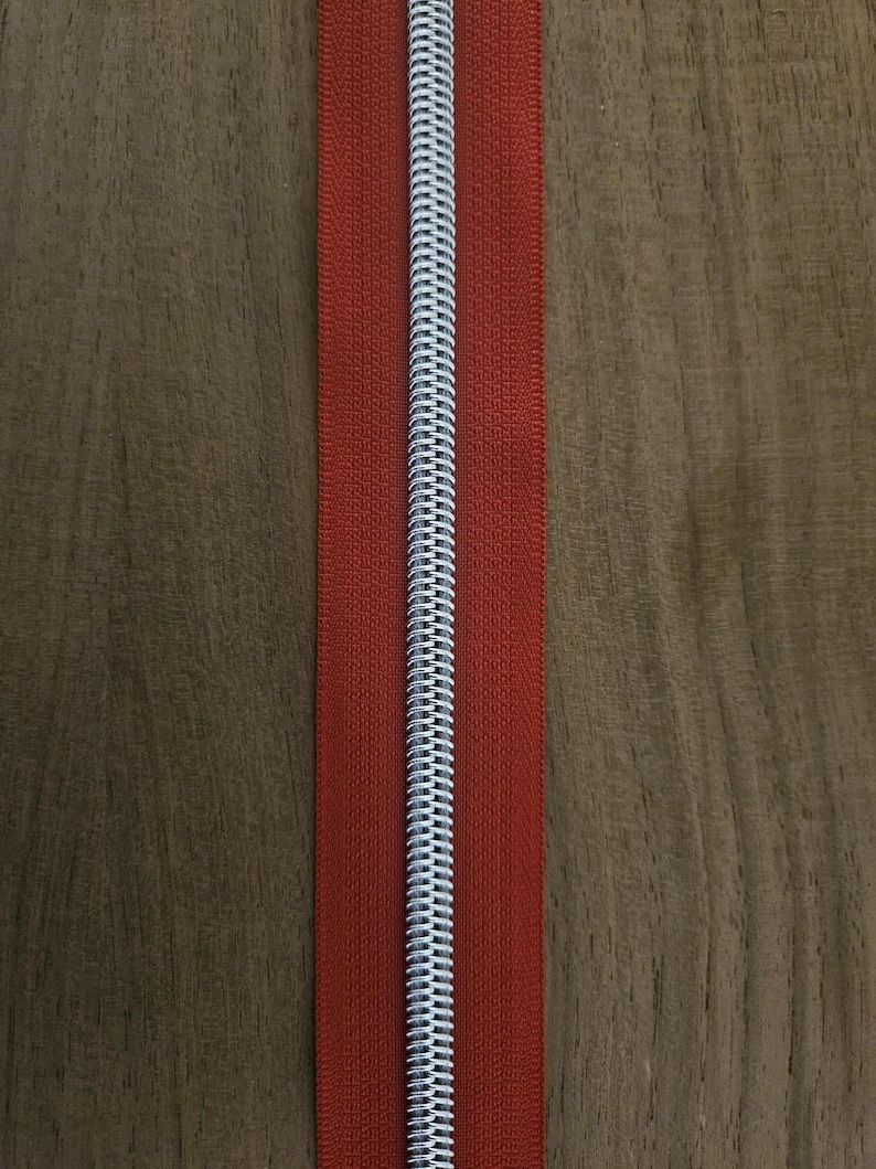 Puede incluir: Primer plano de una cremallera roja con un tirador de metal plateado. La cremallera est&aacute; centrada en una superficie de madera. Los dientes de la cremallera son visibles y la tela es de color rojo liso.