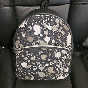 May include: A black leather backpack with a white and gray paint splatter design. The backpack has a zippered pocket on the front and a silver zipper pull.